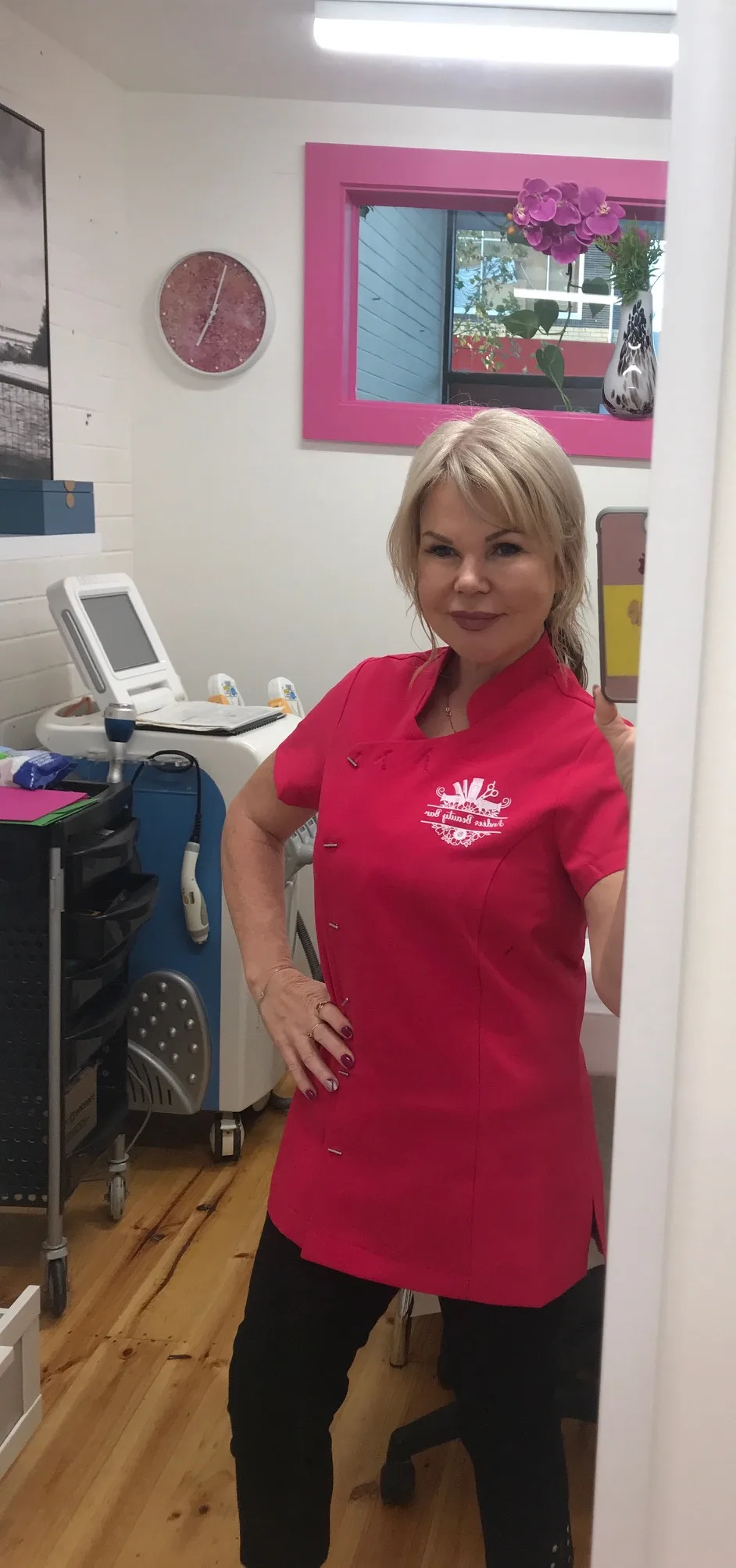 Andée in her hot-pink Andées Beauty Bar tunic — mirror selfie inside a treatment room
